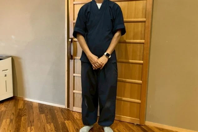 紺色の作務衣(または類似の和装の制服)を着用し、屋内の木目調の床と木製ドアの前で直立している人物。