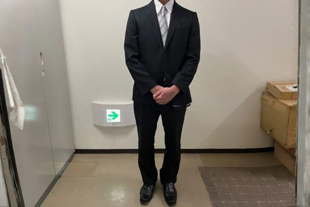 黒いスーツとパンツ、白いシャツにストライプ柄のネクタイを着用した男性スタッフの全身画像。ベル・フロント・ドライバーの制服の画像。