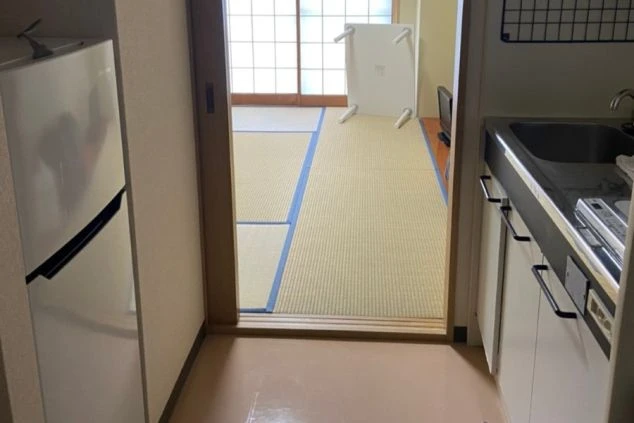 奥に窓がある狭いキッチンで、自然光が差し込んでいます。シンクとコンロがあるカウンターが右側にあり、左側には収納キャビネットがあります。部屋は清潔で整頓されています。