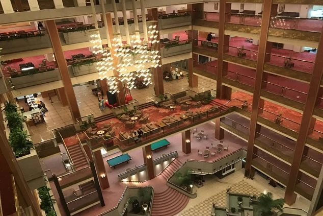 豪華なホテルロビーの鳥瞰図。暖色の照明が空間全体を照らし、豪華な木製家具、装飾的な柱、そして輝くシャンデリアが特徴です。多くの人々がロビー内を歩き回っており、活気と賑わいが感じられます。