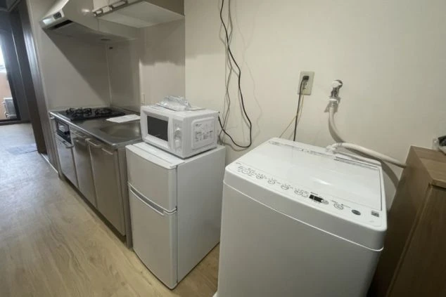 寮のキッチン周り。シンクとコンロが並ぶキッチンカウンター、横に冷蔵庫と電子レンジ、さらに全自動洗濯機が置かれている。寮内の生活設備。