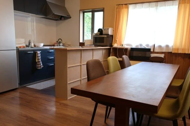 広いダイニングキッチン。茶色のテーブルと椅子が並び、奥に冷蔵庫と黒い調理台がある。