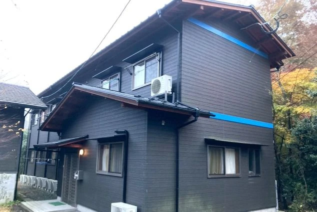 雪が残る中、森に囲まれた黒っぽい二階建ての建物の外観。手前に白い車の一部が見える。