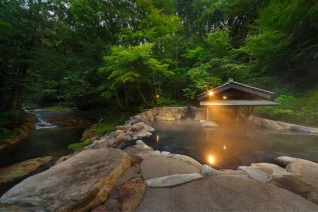 森に囲まれた露天風呂。湯船の傍には屋根付きの休憩所があり、横には小川が流れている。