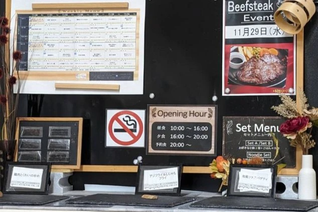 寮の食堂の掲示板。週替わりのメニュー表、営業時間、そしてステーキやセットメニューの告知ポスターなどが貼られ、下の台にはサンプル料理名(アジフライなど)の札が置かれている。