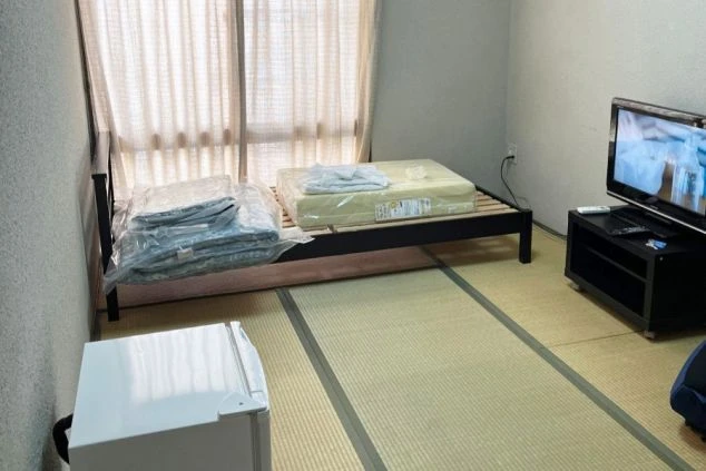 畳敷きの和室個室。すのこタイプのベッドフレームが置かれ、その上には新しい寝具一式が用意されている。室内にはテレビ、小型冷蔵庫、窓には薄手のカーテンがある。