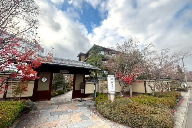冬の柔らかな日差しに照らされた、風格ある和風旅館の入り口。瓦屋根の立派な門と土塀が続き、赤く色づいたもみじや手入れされた植栽が、落ち着いた和の風情を醸し出している。