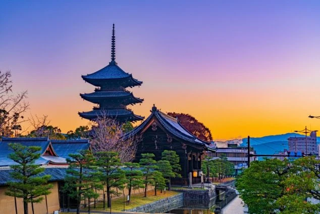 夕暮れ時の美しいグラデーションの空にそびえ立つ、歴史ある五重塔と寺院の建物。手前には手入れされた松の木が並び、遠くの山々や街並みと調和した京都らしい風景。