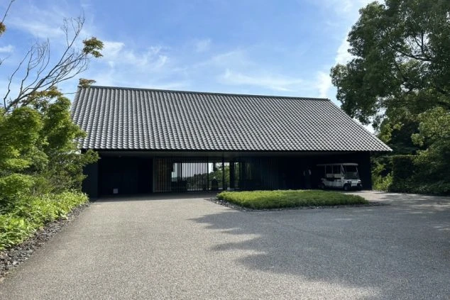 建物の正面。大きな瓦屋根と黒い壁のモダンな和風建築で、全面ガラス戸の向こうに室内が見える。手前は手入れされた芝生で、飛び石がある。