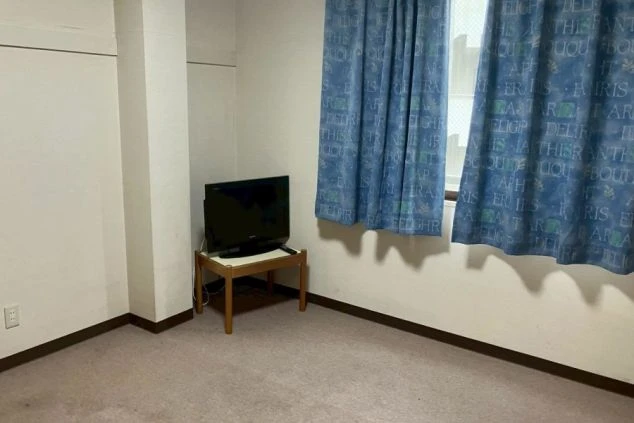 青いカーテンの窓際におかれた、テレビとテレビ台のあるシンプルな洋室。