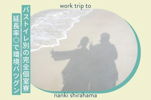 澄み渡る青空の下、白い砂浜に設置された藁ぶき屋根のパラソル。遠くに海岸沿いの街並みが見える。