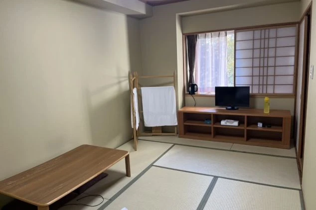 寮の個室。畳敷きの和室で、窓の外には緑の山と湖または川が見える。テレビ、ケトル、木製のテレビ台やタオル掛けなどが備え付けられている。