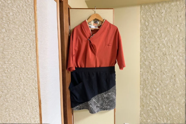 ハンガーに掛けられた、吉祥のレストランスタッフの制服。朱色（またはオレンジ色）の和風上着と、裾に波柄がデザインされた紺色の巻きスカートまたはエプロン。