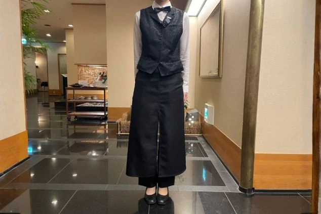 黒いベストとボウタイ、白いシャツを着用した従業員(人物は胸から下のみ)が、ホテルのロビーで直立している。