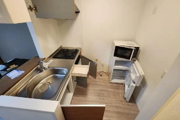 白い壁と木目調の床の部屋にある、流し台、コンロ、電子レンジ、小型冷蔵庫。