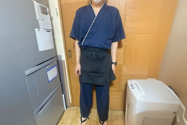 紺色の作務衣と黒いエプロンを着用した男性スタッフ。旅館やホテルでのレストラン業務や駐車場係など、リゾートバイトで和風の制服に興味がある方に訴求する画像。