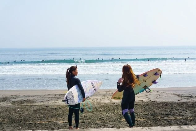 砂浜でサーフボードを抱え、海を見つめる2人の女性サーファー。リゾートバイトでサーフィンを楽しむ休日のイメージ。