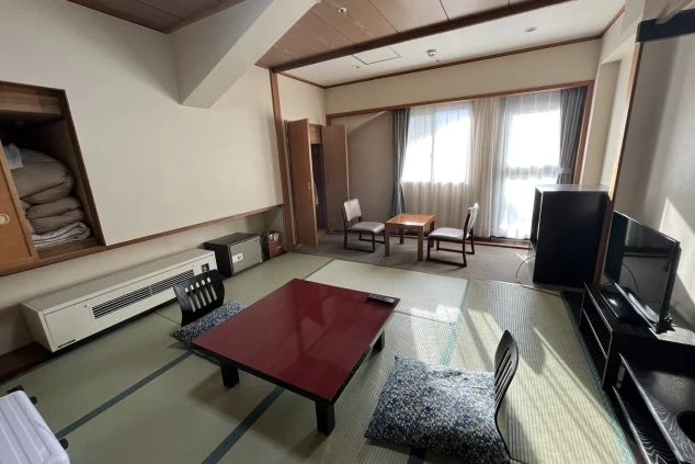 畳敷きのスペースがある和室風の寮の部屋。部屋にはテレビ、こたつテーブル、座布団、そして二つの椅子が置かれている。