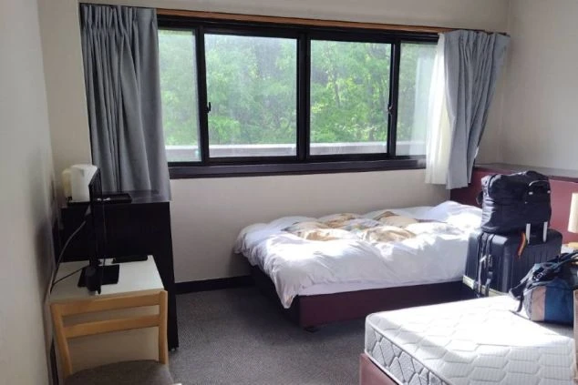 窓から緑が見える寮の部屋。二つのマットレスベッドと、テレビや電気ケトルが置かれたテーブルがある。