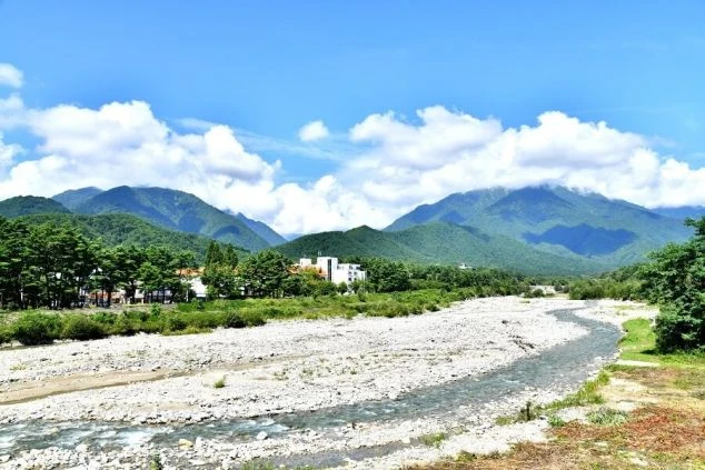 晴れた日の山と川の景色。青々とした山々を背景に、石がごろごろした河原と小川が広がり、遠景には白い建物がいくつか見える。空には白い雲が浮かんでいる。