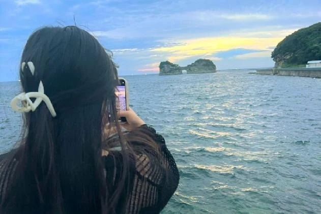 夕暮れ時の海を背景に、中央に穴が開いた特徴的な形の島（円月島）をスマートフォンで撮影している女性の後ろ姿。