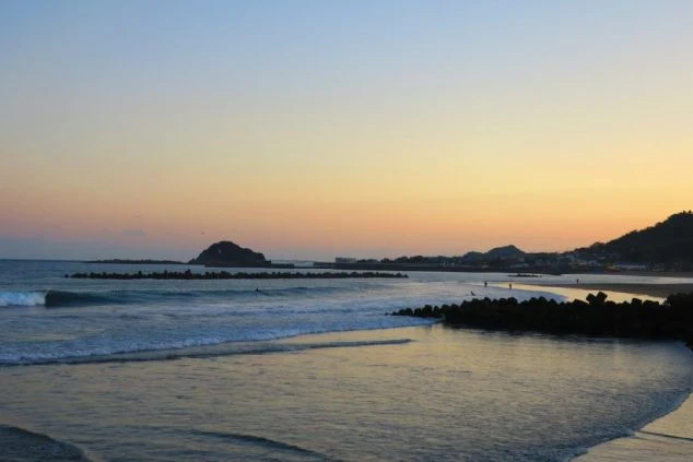 オレンジ色のグラデーションが広がる空と、静かに打ち寄せる波が印象的な夕方のビーチ。サーフィンや散歩を楽しめるリゾートバイトのイメージ画像。