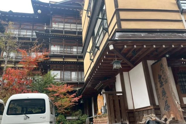 外観。左奥には千と千尋の神隠しを彷彿とさせる、複雑な構造と瓦屋根を持つ伝統的な木造建築の建物がそびえ立ち、手前には比較的新しい黄土色の棟が並んでいる。手前には紅葉した木々や白い軽トラックが見える。