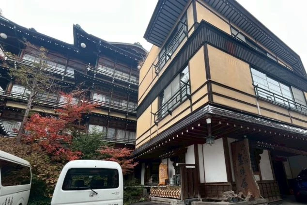 外観。左奥には千と千尋の神隠しを彷彿とさせる、複雑な構造と瓦屋根を持つ伝統的な木造建築の建物がそびえ立ち、手前には比較的新しい黄土色の棟が並んでいる。手前には紅葉した木々や白い軽トラックが見える。