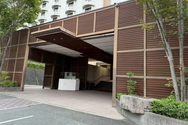 木目調のルーバーで覆われたモダンな車寄せの先に、屋内へ続く階段が見える。奥には客室のバルコニーが連なる高層棟の一部が写っている。