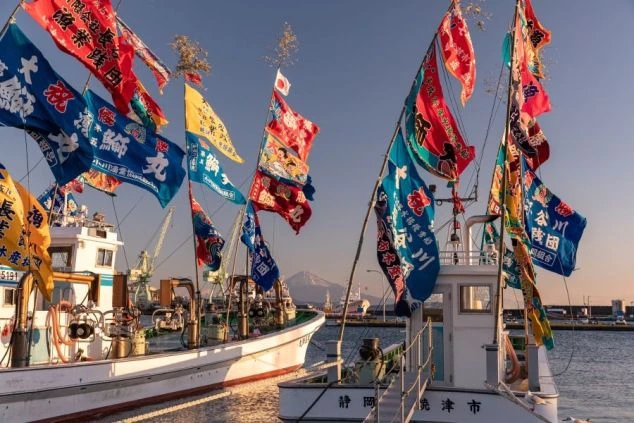 大漁旗を掲げた漁船が並ぶ焼津港。晴れた空の下、遠くに富士山が見え、活気ある港の様子が伝わる。