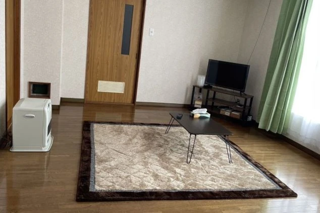 フローリングの床のリビングルーム。中央にはラグとローテーブルが置かれ、テレビ、ファンヒーター、そしてドアが見える。