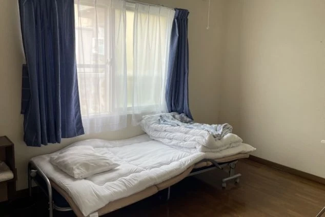 窓に青いカーテンがかかったシンプルな部屋。床は木製で、折りたたみ式のベッドに布団が敷かれている。