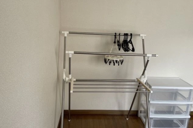 部屋の隅にある物干しラック。服がいくつか掛かっており、横にはプラスチック製の引き出しがある。