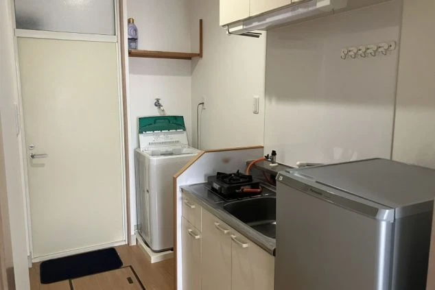 寮の個室内にあるミニキッチンと洗濯機スペースの画像。ガスコンロ、冷蔵庫、電子レンジが揃い、自炊派にも嬉しい充実の生活環境。