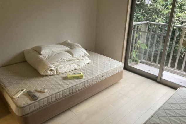 採光の良い清潔感のある寮の部屋。窓際にはバルコニーが見え、室内にはマットレスと布団、ティッシュ、テレビのリモコンが置かれており、リゾートバイトで生活する寮をイメージさせる