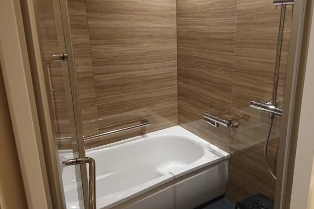 モダンなバスルームで、白いバスタブとシャワーが備わっています。壁は木目調のタイル張りで、床には暗い色のマットが敷かれています。