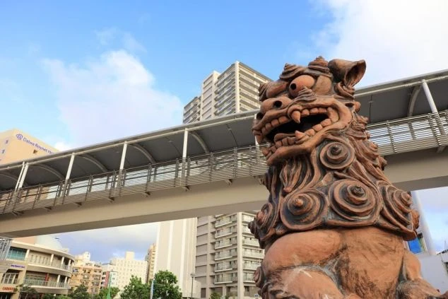 青い空の下、茶色いシーサーの像がクローズアップで写っています。背景には、木々が生い茂る丘と、その奥に建物が見えます。