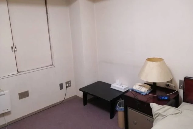 寮の個室。ベッドサイドにランプ、電話、時計、小さな机とゴミ箱が映る。