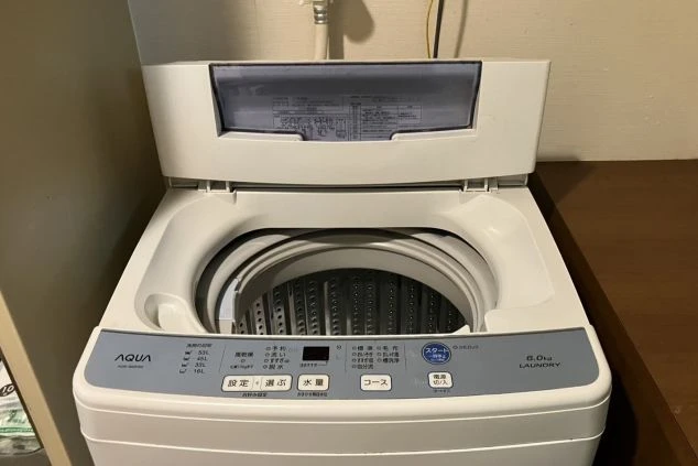 寮に設置されている洗濯機の画像。容量6.0kgの家庭用洗濯機で、無料で使える共同設備であることを示唆し、生活費を抑えられるメリットを伝えている。