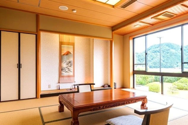 リゾートバイトの勤務先の旅館の畳敷きの広々とした和室。大きな窓からは緑豊かな景色が見え、奥には掛け軸と床の間がある。