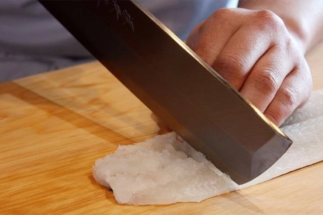 まな板の上で、大きな包丁を使って魚の切り身を薄くスライスしている人の手元を写したクローズアップ画像です。包丁は銀色の刃で、黒い柄が付いています