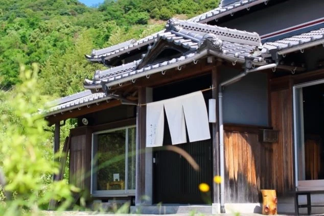 古い伝統的な日本家屋のクローズアップで、木製の引き戸と、その上に吊るされた白い暖簾が特徴です。暖簾には日本の文字が縦書きで書かれています。