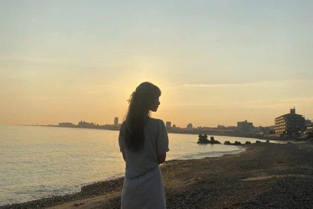 水辺に立つ人物のシルエットが写っている夕暮れの屋外写真です。人物は画面の中央やや左に位置し、横向きで、長い髪をしています。夕日が地平線近くにあり、空はオレンジと黄色の暖かい色合いに染まっています。