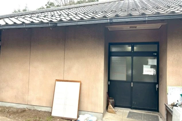 瓦屋根とベージュの外壁の平屋建ての建物。黒いサッシの引き戸が入口となっており、軒下には竹ぼうきや立てかけられた障子が置かれている。