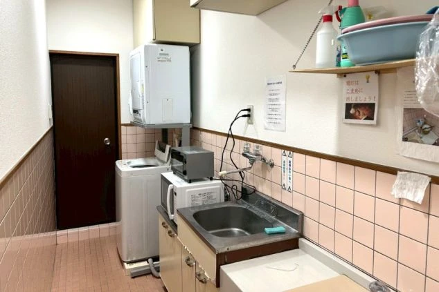 共用の水回りスペース。ステンレスの流し台の横に、洗濯機とその上に乾燥機、電子レンジ、トースターなどの家電がコンパクトに配置されている。