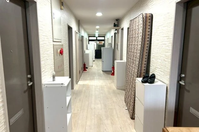 白い壁と木目調の床が続くアパートの共用廊下。各部屋のドアが並び、廊下には冷蔵庫や棚、立てかけられたマットレスが置かれている。