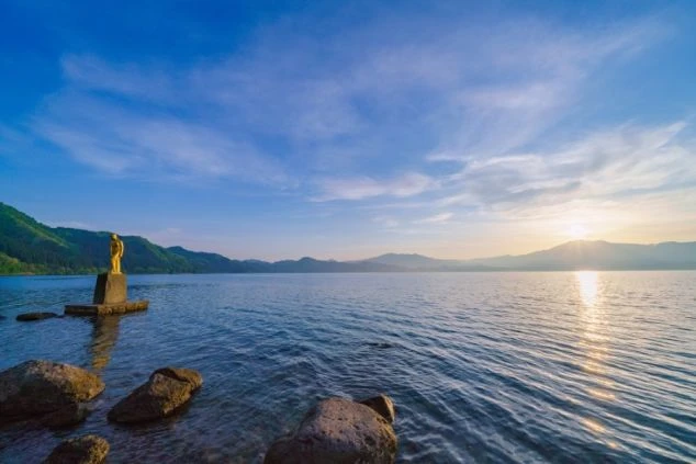 田沢湖に立つ黄金のたつこ像。夕焼けの湖面に光が映る幻想的なイメージ画像。リゾートバイトの休日を満喫。
