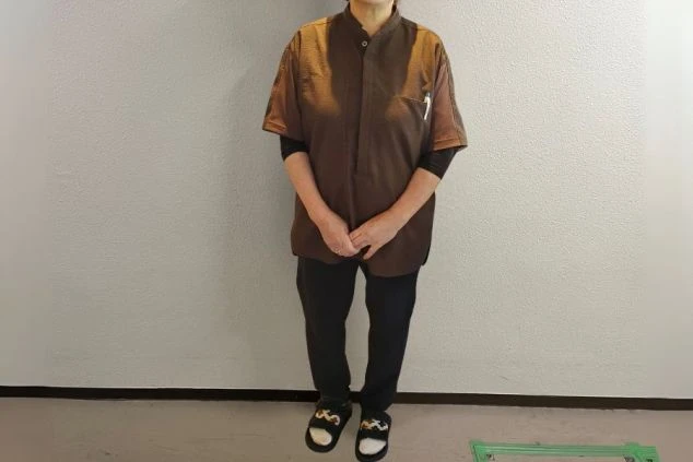 濃い茶色の作務衣風トップスに黒いパンツ、サンダルを着用した男性スタッフの全身画像。裏方やカジュアルな接客に適した動きやすい服装である。リゾートバイトの仕事内容を表す、接客全般スタッフの制服の画像。