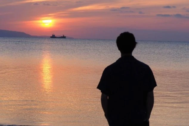 海に沈む夕日を眺める男性の後ろ姿。オレンジ色に染まる空と、きらめく水面が、エモーショナルな雰囲気を醸し出している。
