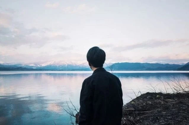 湖畔で雪化粧をした山々を眺めている男性の後ろ姿の画像。穏やかな水面と空が広がり、リラックスした雰囲気が漂う。リゾートバイトの休日の過ごし方や自然の景色を表す画像。